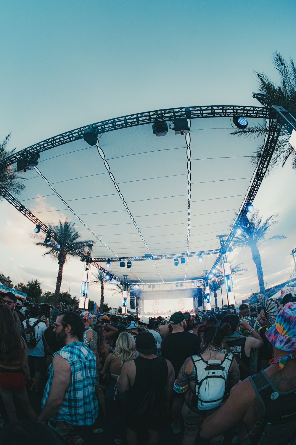 Crowd at outdoor music festival stage with palm trees and truss lighting at dusk.