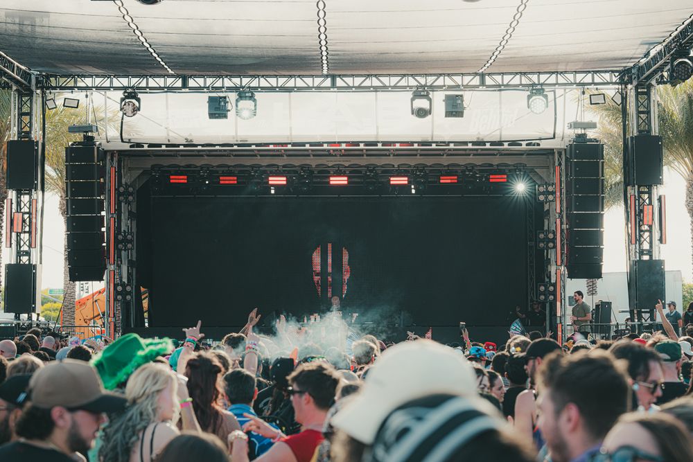 Large crowd at an outdoor music festival with stage lights and performers visible on a dark stage.