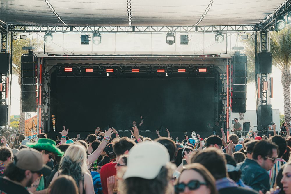 Large crowd of festival-goers with raised hands facing a lit outdoor concert stage at a music festival.