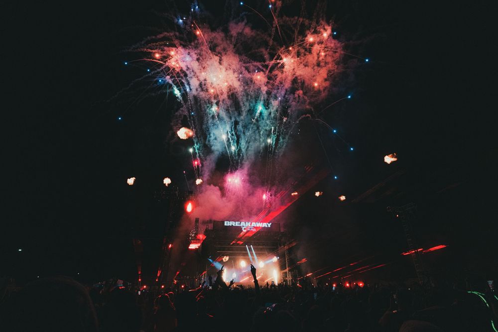 Colorful fireworks explode over a crowded outdoor music festival stage at night.