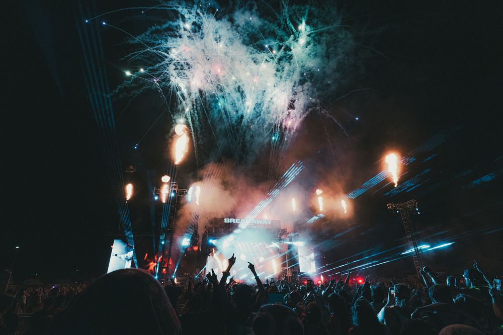 Crowd at outdoor concert with colorful fireworks and stage lights illuminating the night sky.