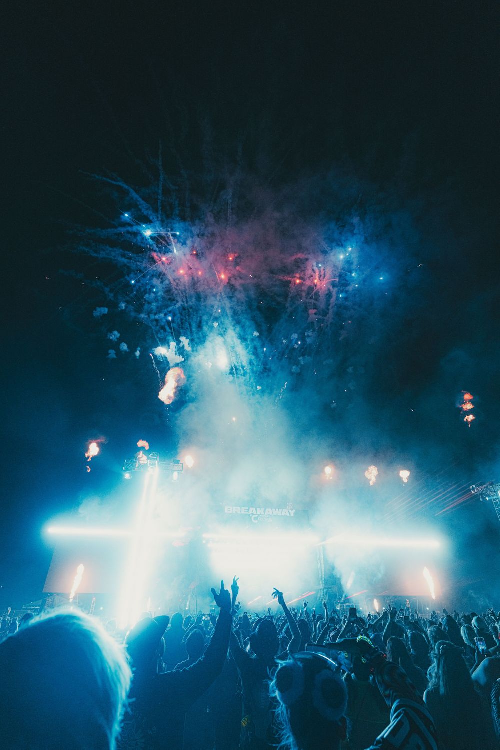 Crowd cheering at an outdoor music festival with blue stage lights and colorful fireworks exploding in the night sky.