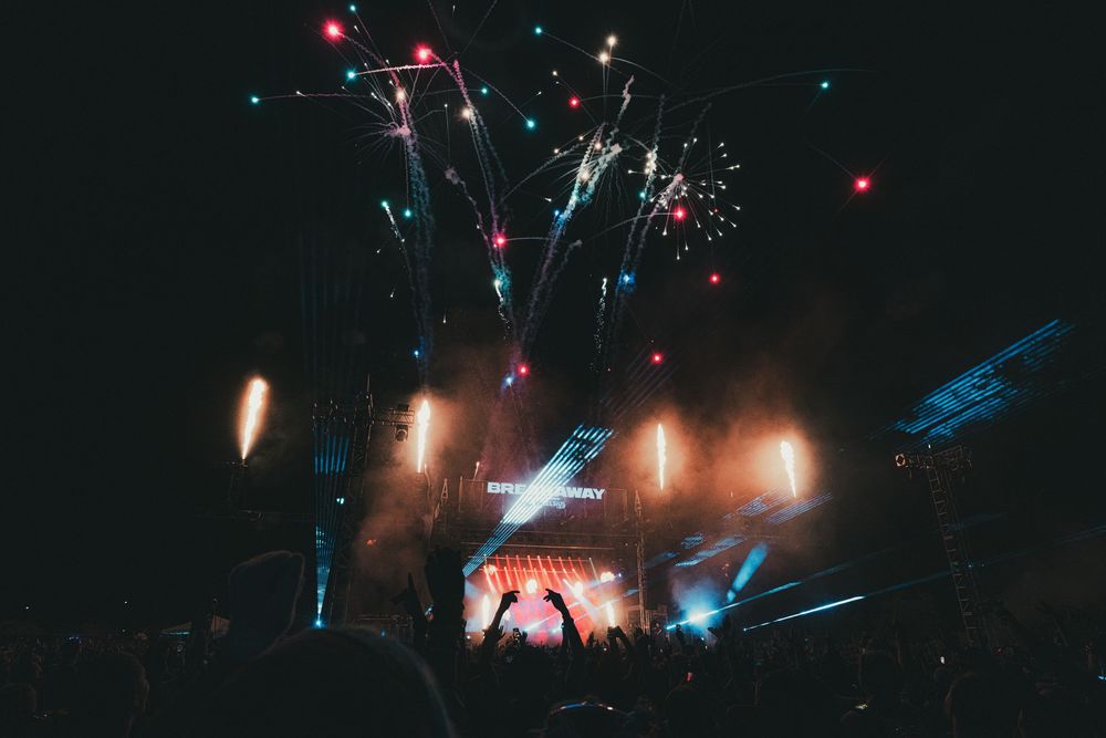 Crowd at outdoor concert with colorful fireworks and stage lights illuminating the night sky.