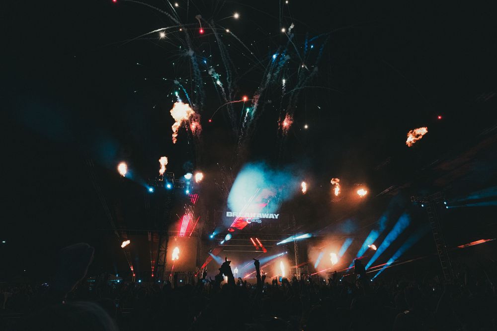 Crowd at an outdoor music festival with colorful fireworks and stage lights illuminating the night sky.
