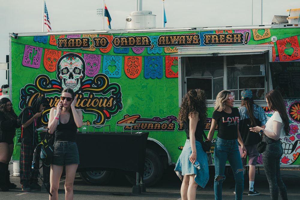 Colorful green food truck with skull art serving fresh made-to-order tacos, with customers gathered outside at a street fair.