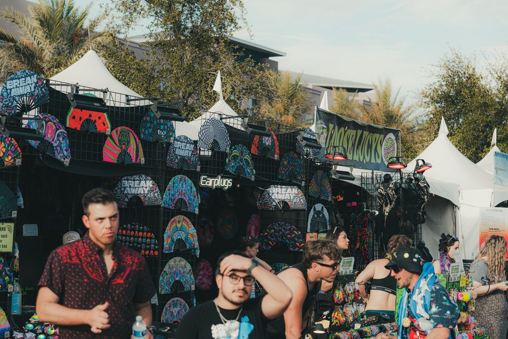 Colorful vendor stalls selling bags and accessories at an outdoor festival market with palm trees in the background.