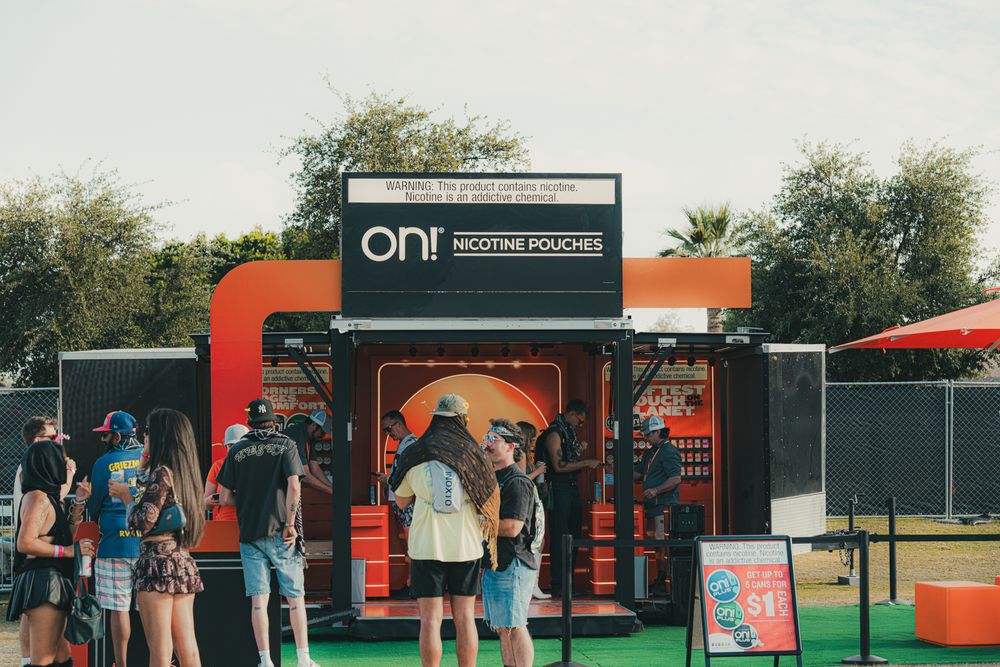 People gathered at an ON! nicotine pouches branded orange festival booth outdoors on a sunny day.