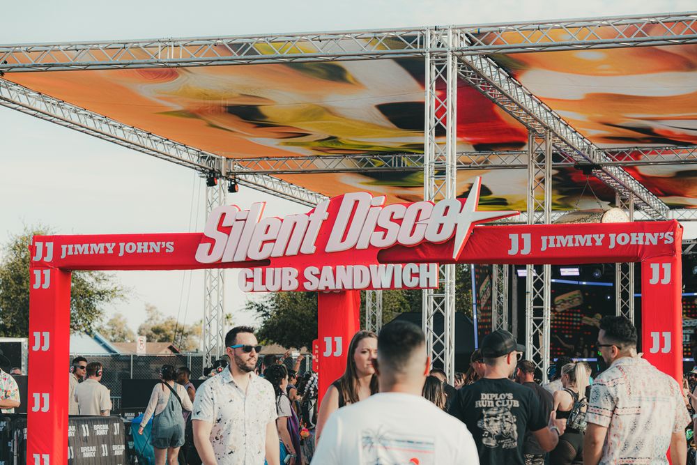Crowd gathered under a Jimmy John's Silent Disco Club Sandwich festival stage with colorful truss lighting.