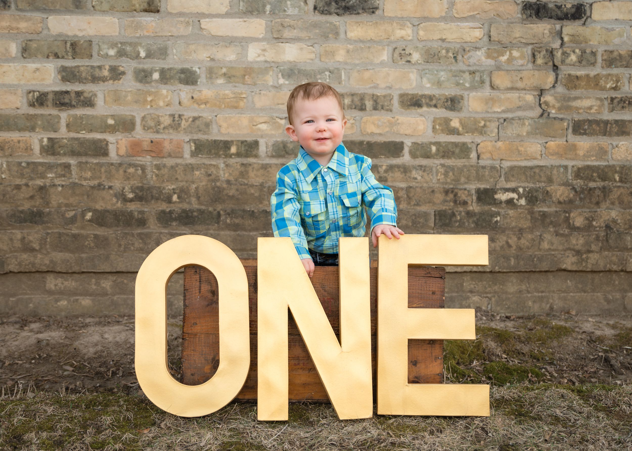 Smiling toddler boy in plaid shirt posing with large wooden ONE letters against brick wall for first birthday photo.
