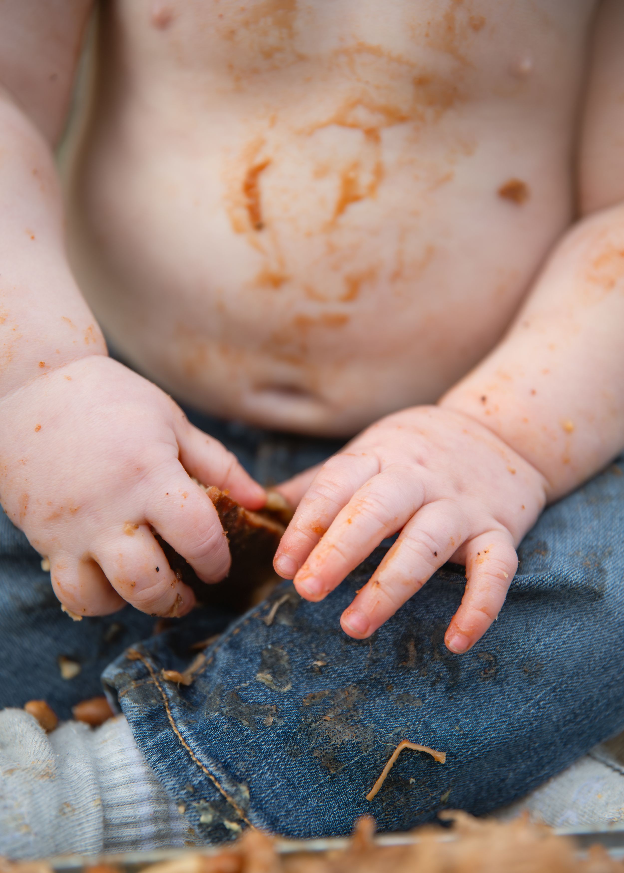 Close-up of messy baby hands covered in cake smash frosting over blue jeans.