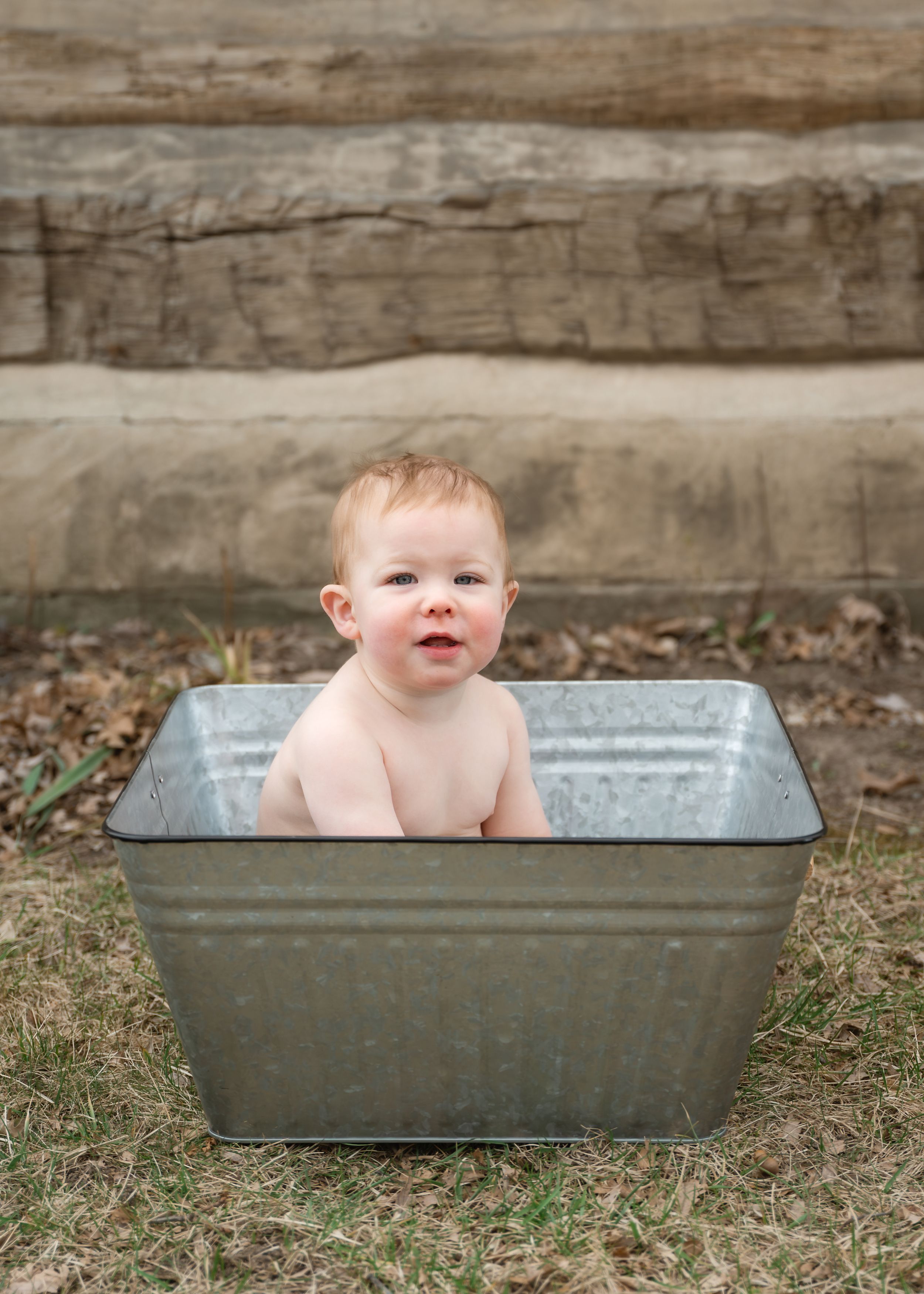 Smiling baby with red hair sits in vintage galvanized metal tub outdoors on grass.