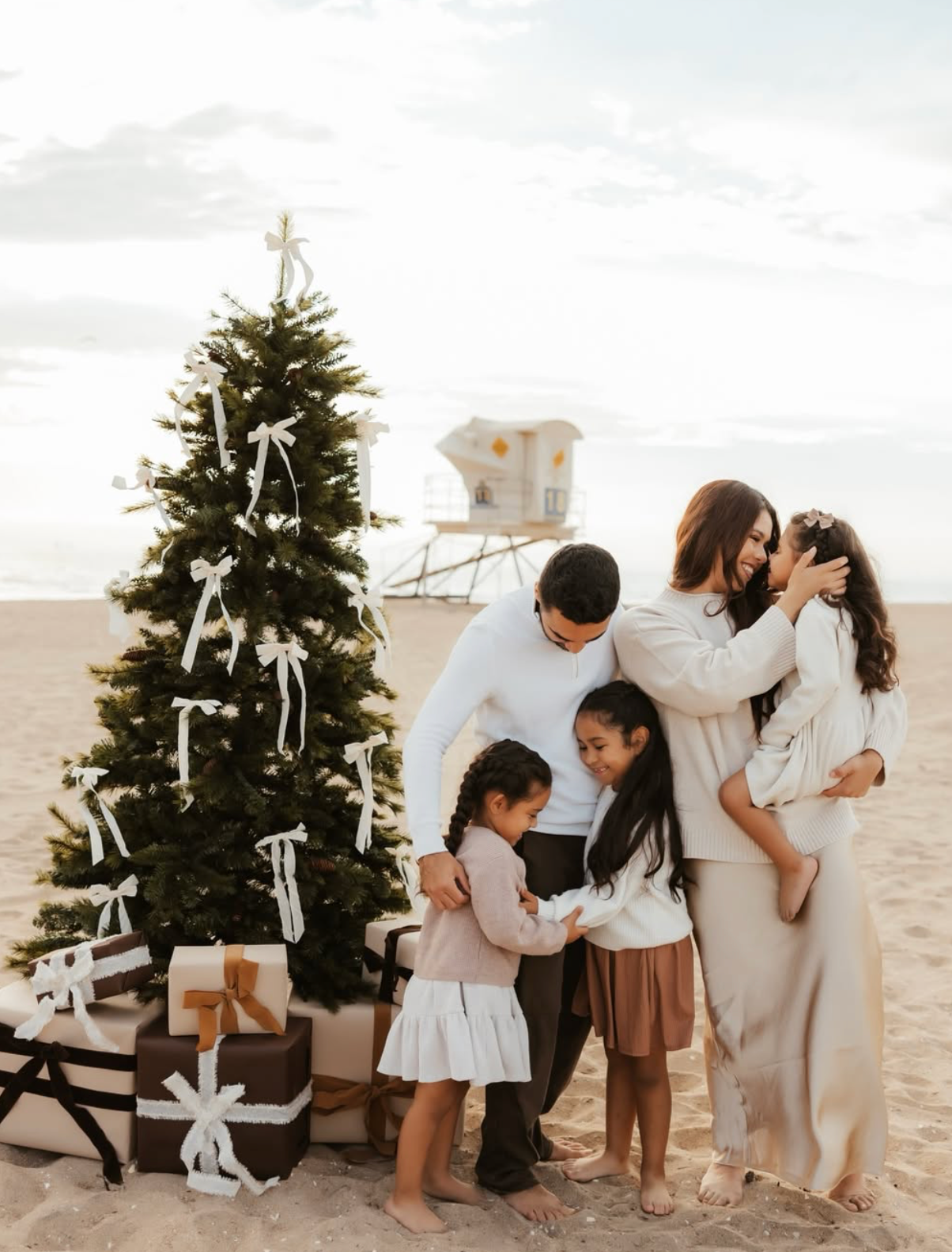 Family gathering around a decorated Christmas tree with gifts on a sandy beach, lifeguard tower in background.