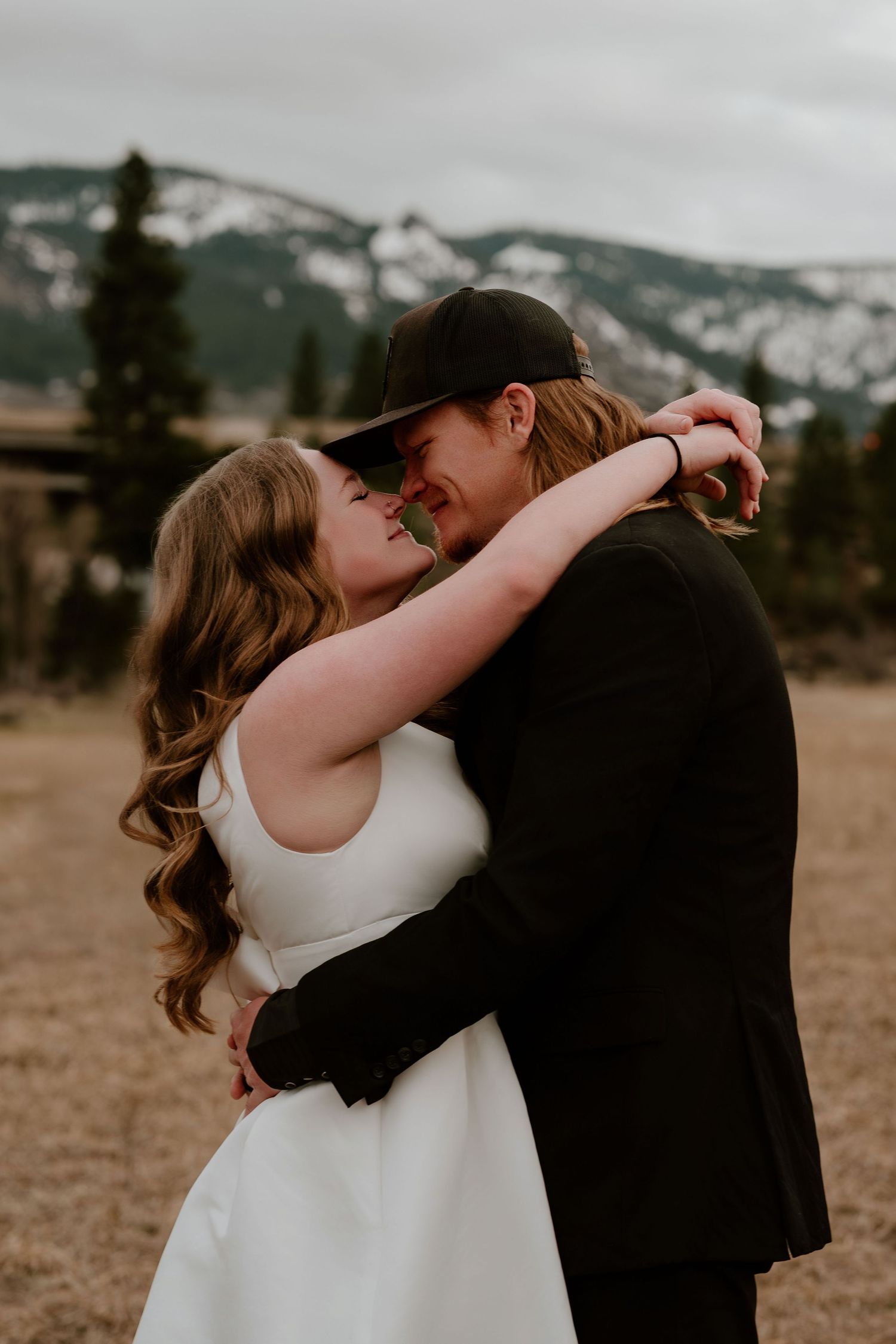 Bride and groom embracing outdoors with snow-capped mountains in background, groom wearing black suit and cap.