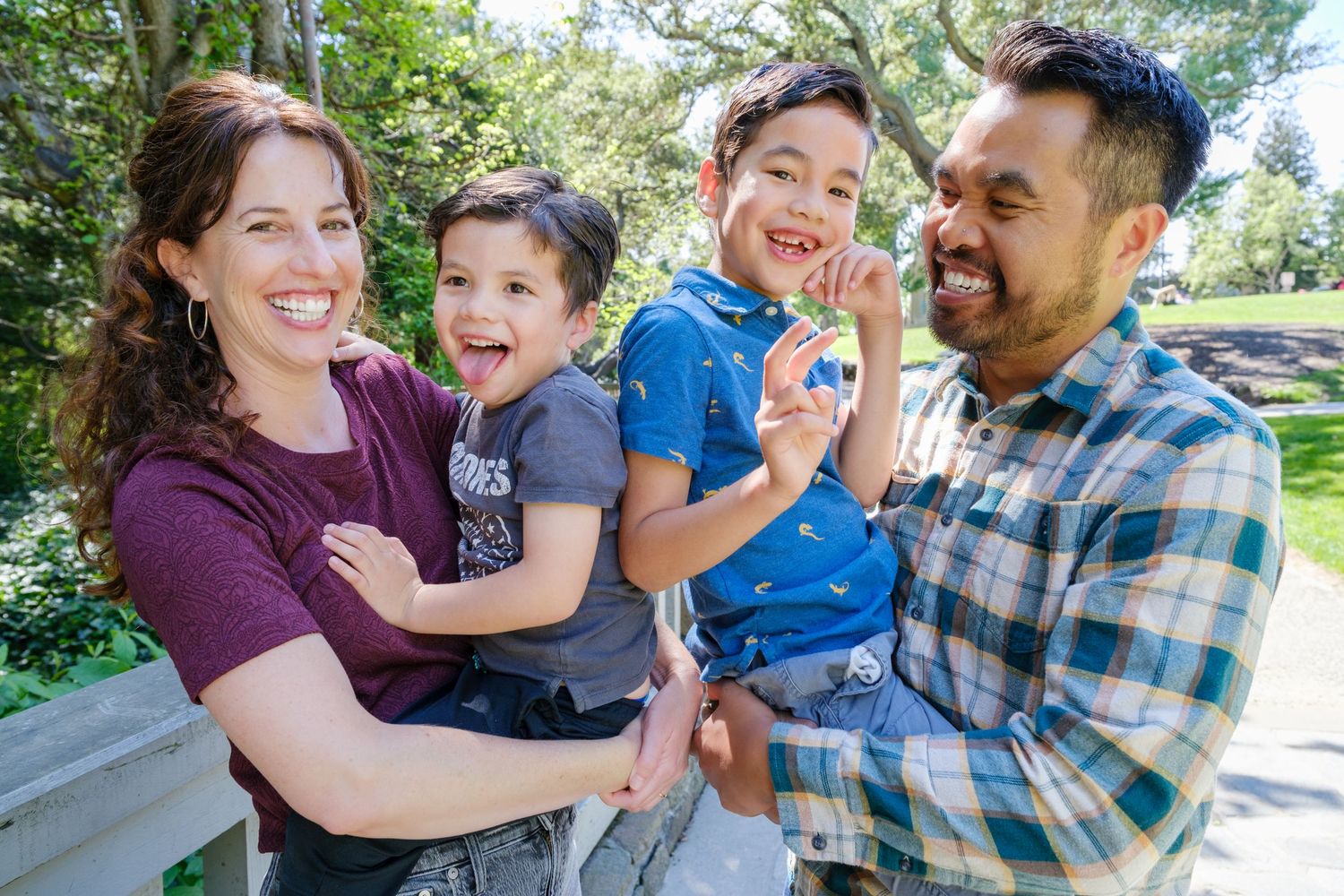 Happy family of four smiling outdoors in a park, parents holding two young boys on a sunny day.
