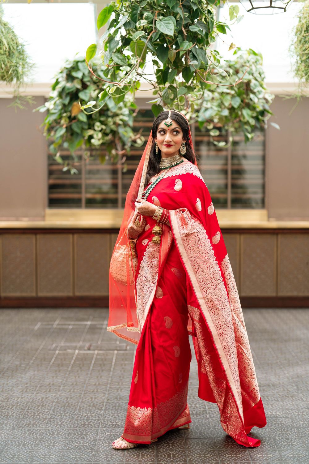 Woman in red Banarasi saree with gold embroidery and bridal jewelry poses elegantly indoors.