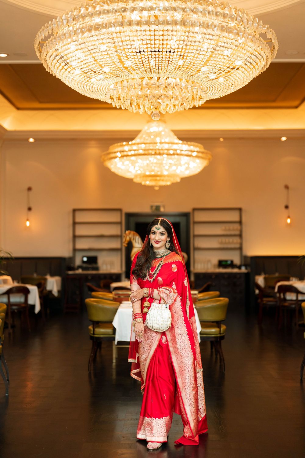 Indian bride in red saree with gold jewelry poses under crystal chandeliers in elegant banquet hall.