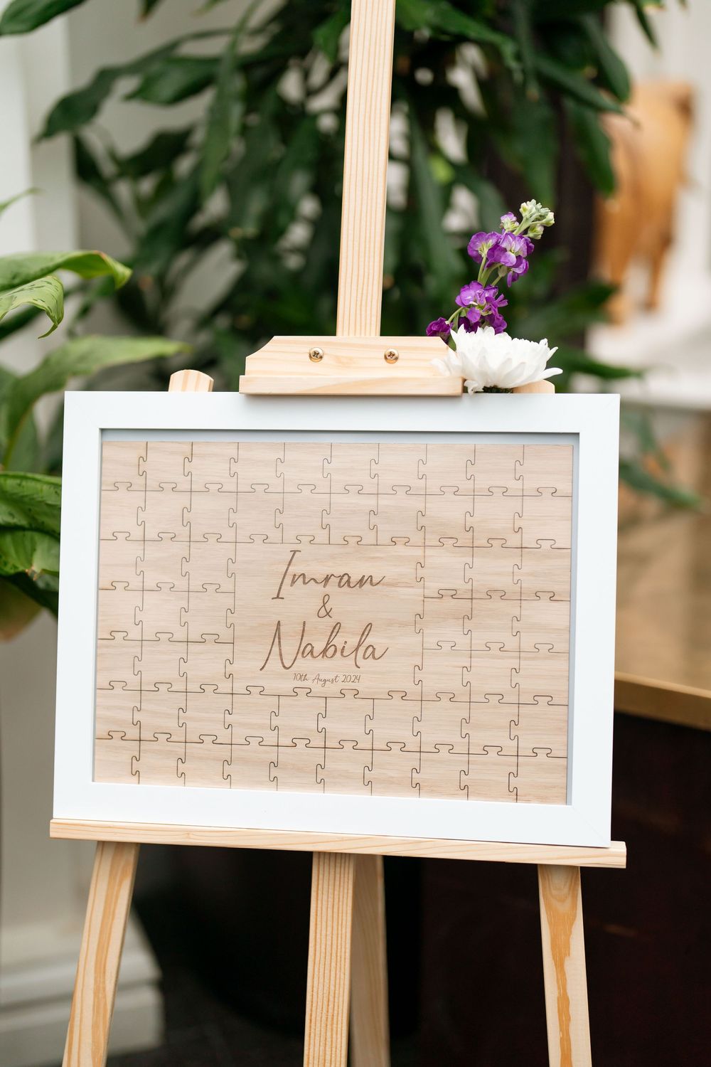 Wooden puzzle guest book on easel with engraved names Imran and Nabila, decorated with purple and white flowers.