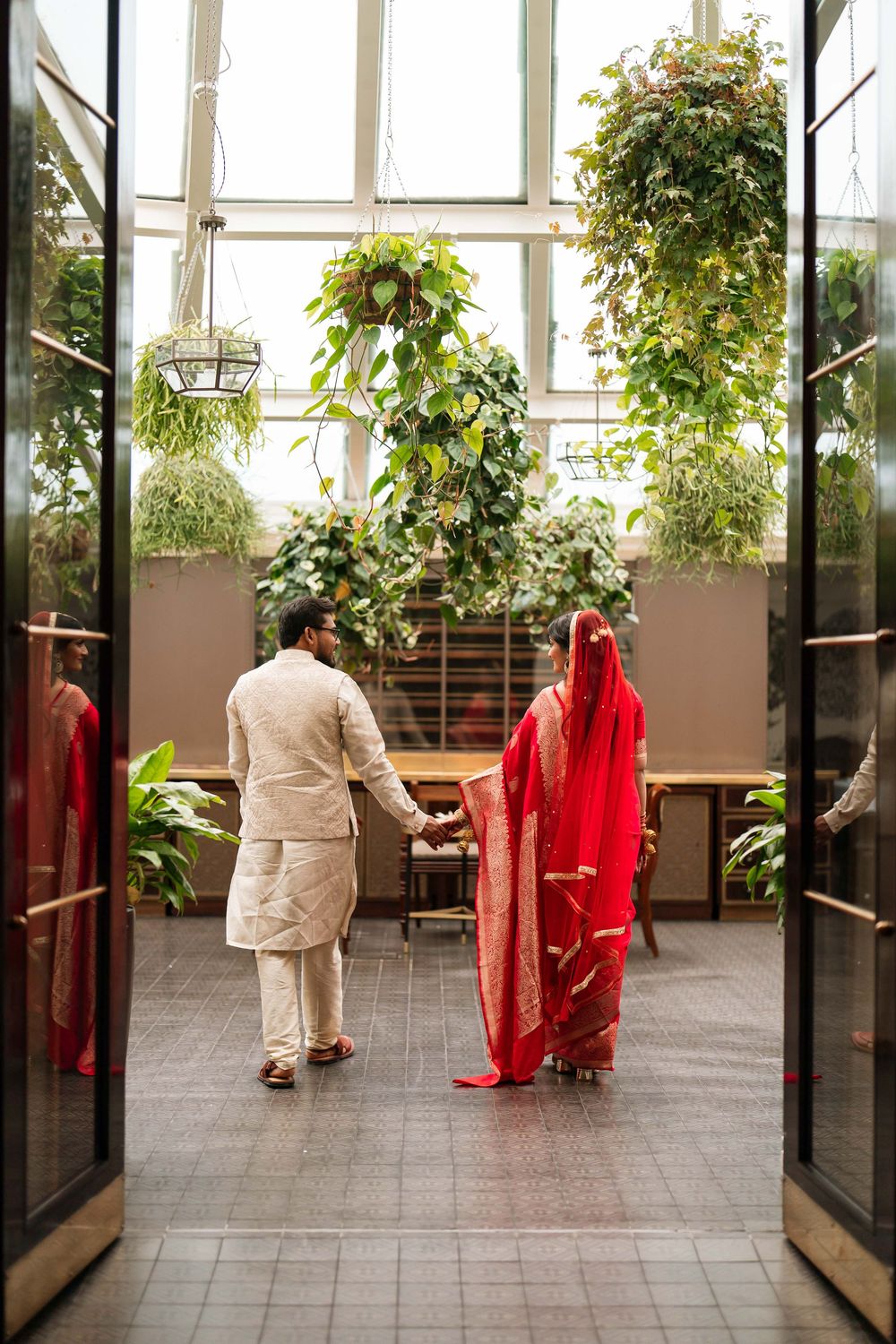 Indian couple in traditional wedding attire holding hands in a lush greenhouse venue, bride in red saree, groom in cream sherwani.