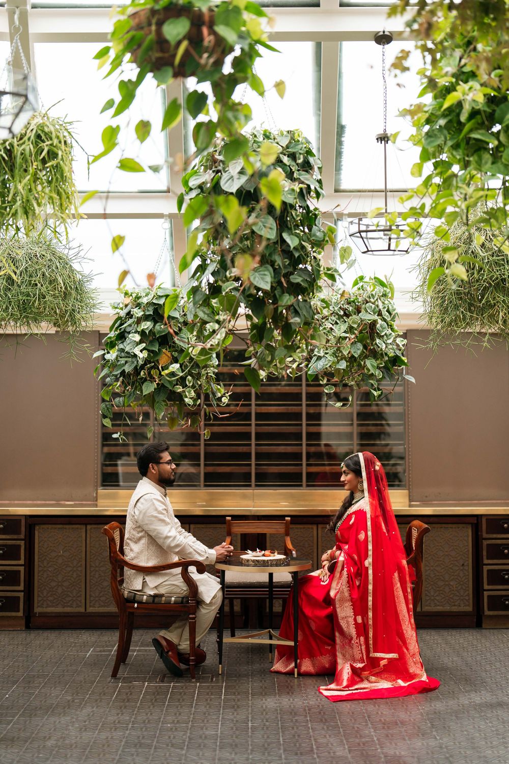 Couple in traditional Indian wedding attire sitting at a table surrounded by lush hanging plants in an elegant indoor setting.