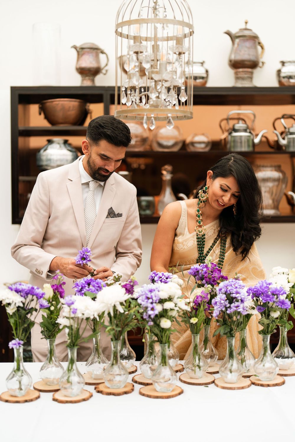 Couple arranging purple and white flowers in glass vases on a white table at an elegant wedding venue.