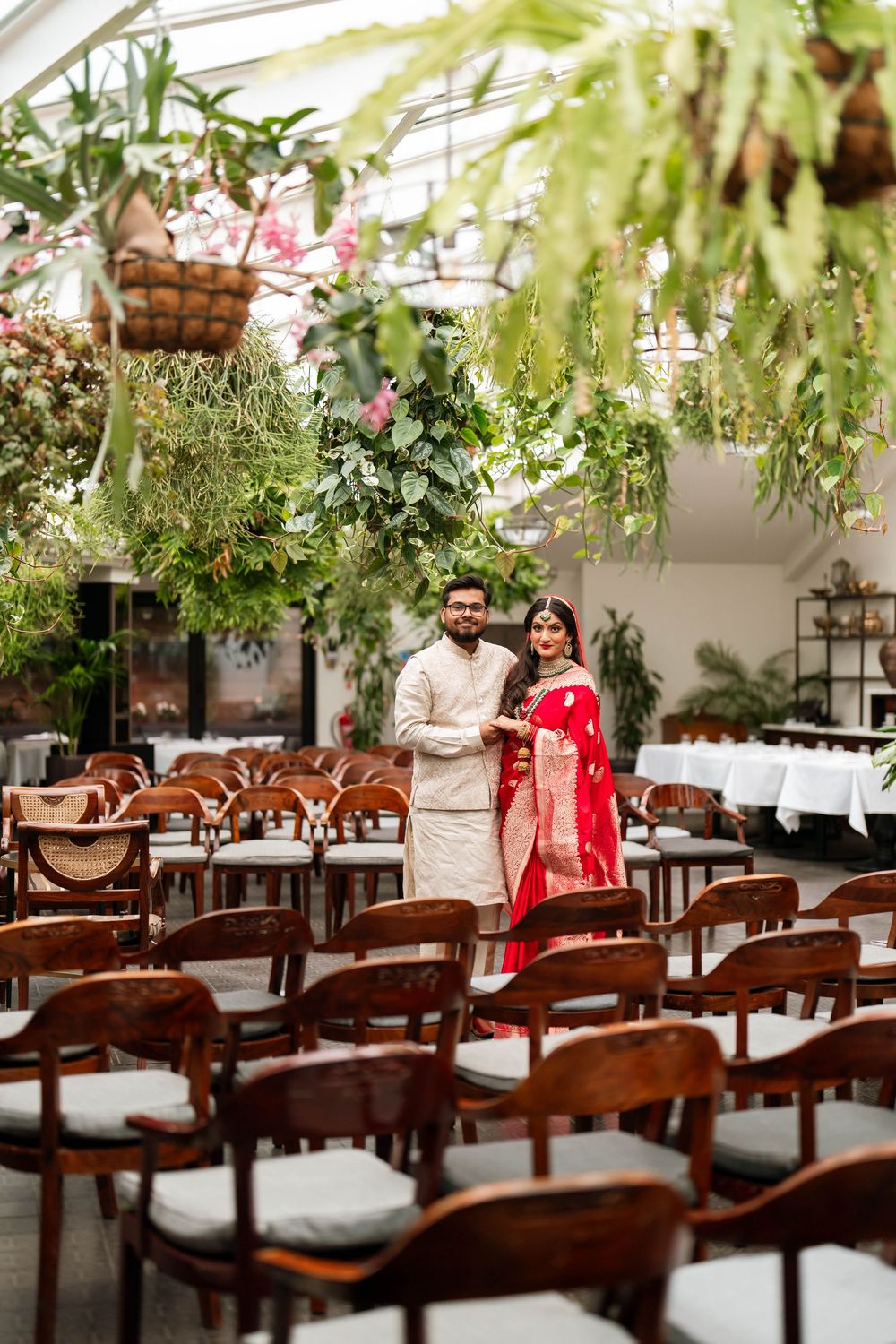 Indian couple in traditional wedding attire standing among wooden chairs in a greenhouse venue with lush hanging plants.