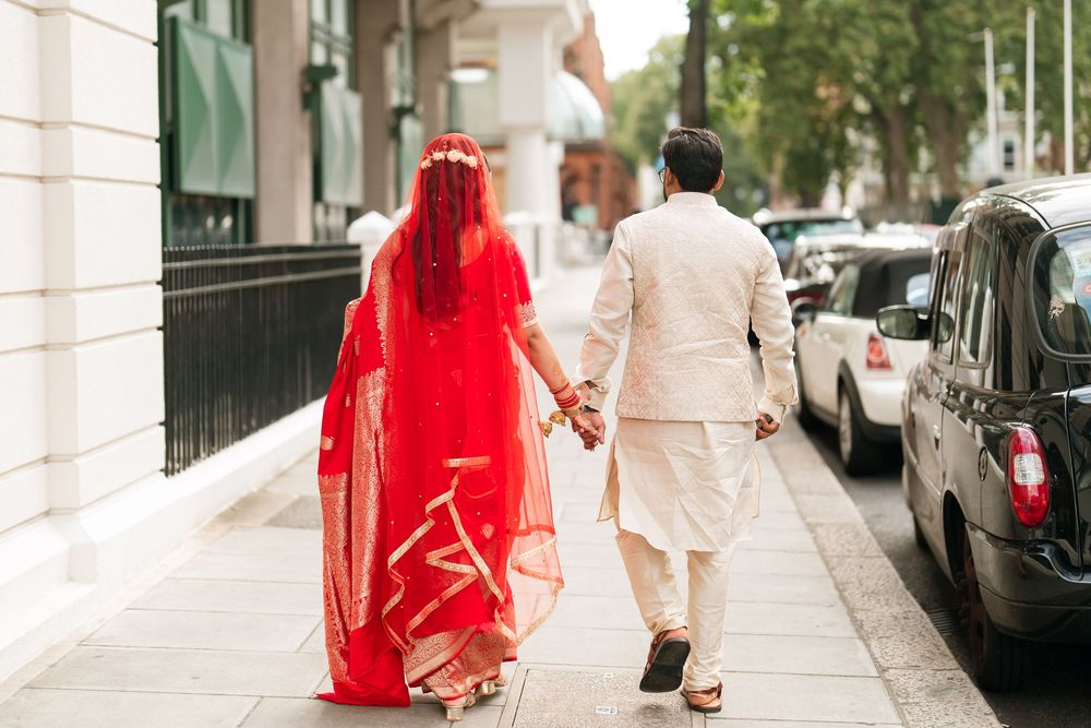 Couple in traditional South Asian wedding attire holding hands walking on a city sidewalk, bride in red dupatta.