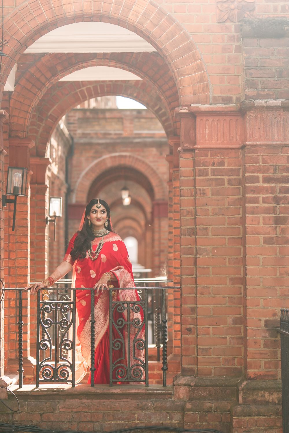 Woman in red saree poses gracefully on ornate iron railing amid red brick arched corridor.
