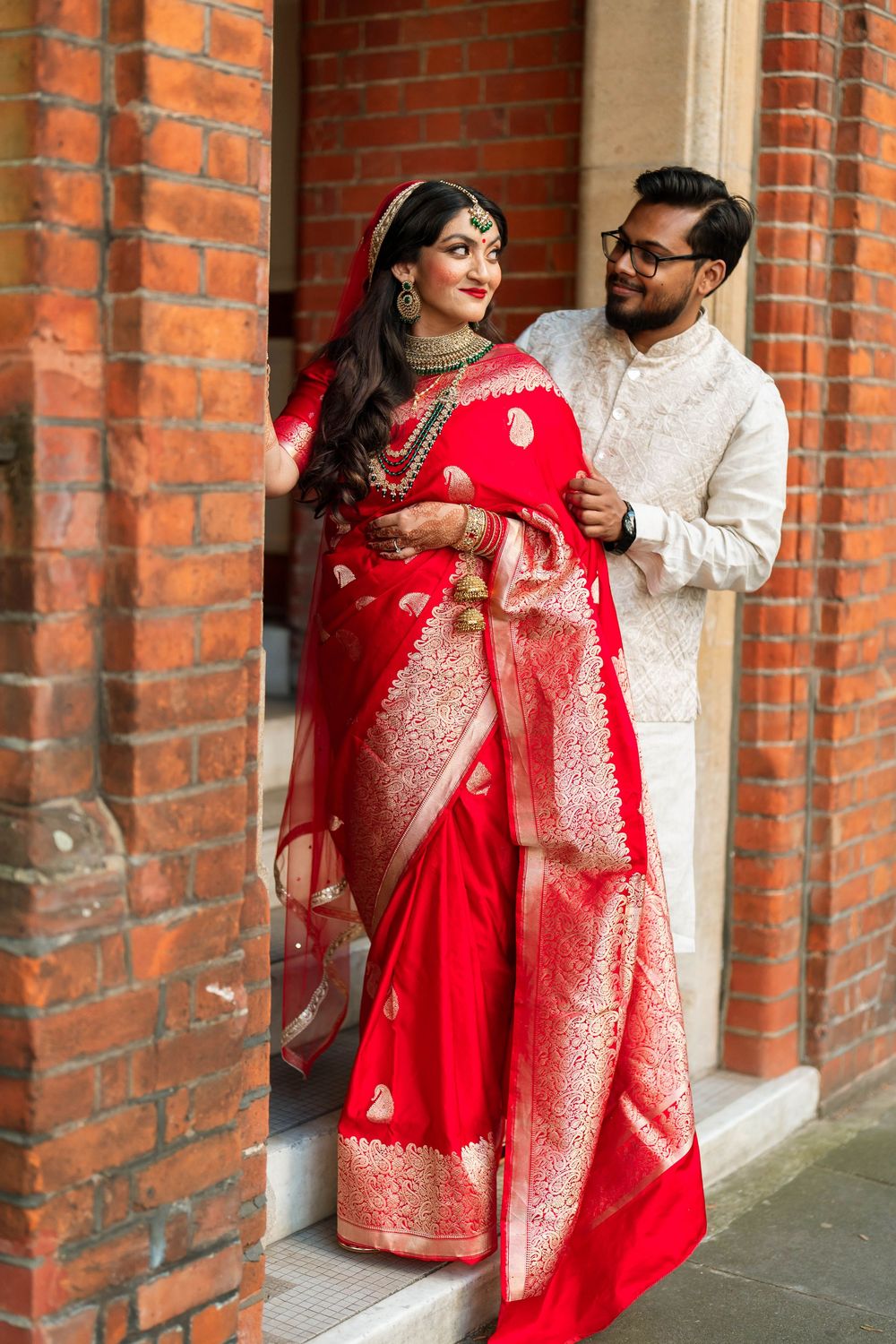 Indian bride in red Banarasi saree with groom in white sherwani posing at brick building entrance.