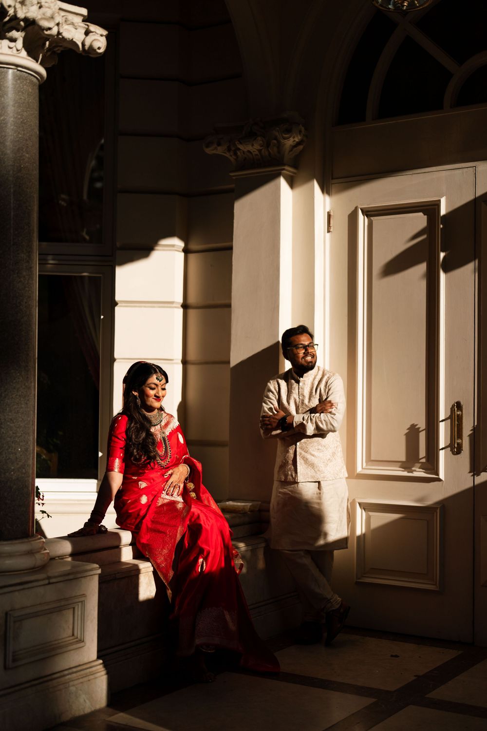 Indian bride in red saree and groom in cream kurta posing at elegant architectural venue with dramatic lighting.