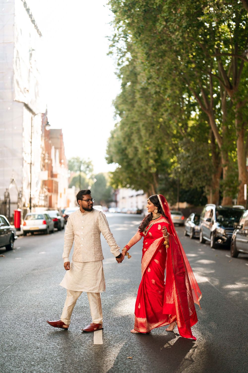 Indian bride in red saree and groom in cream sherwani hold hands walking on a tree-lined street.