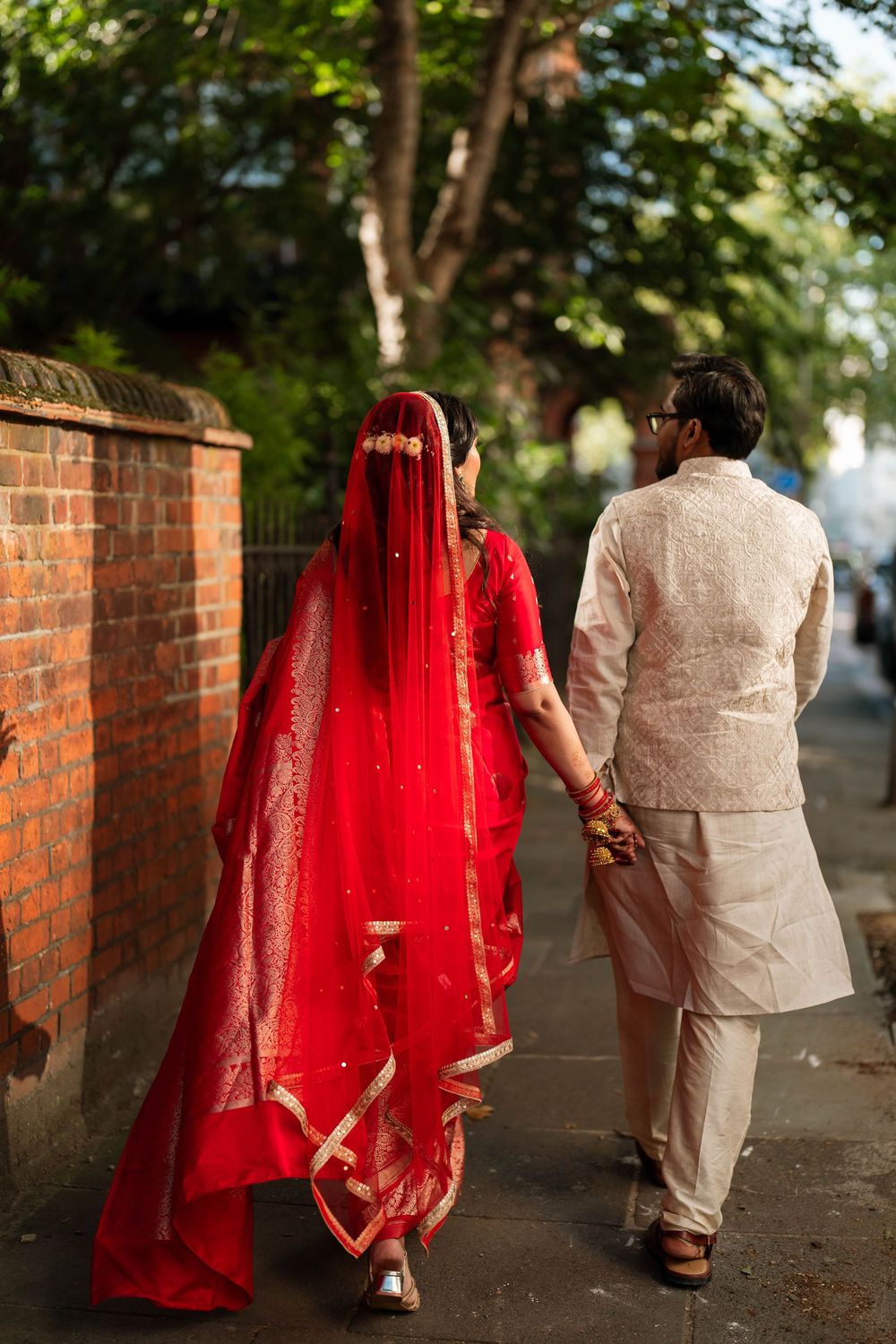 Indian bride in red saree and groom in cream kurta walking hand in hand along a brick wall path.