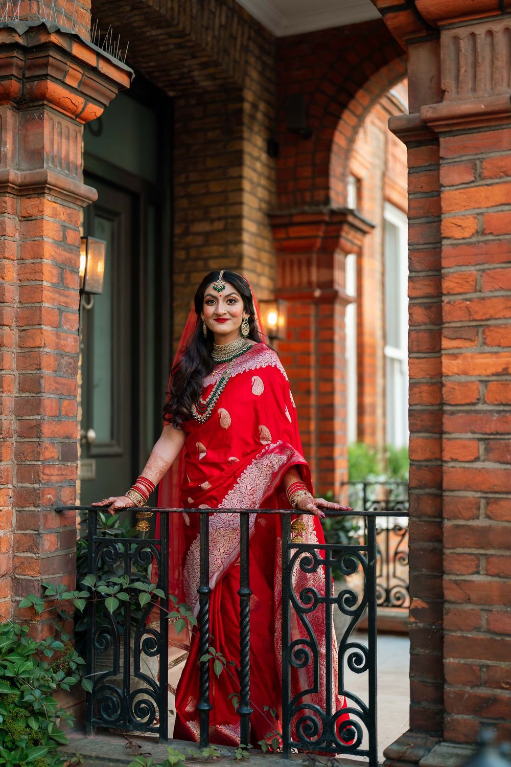Beautiful Indian bride in red Banarasi saree standing on ornate balcony of brick building smiling.