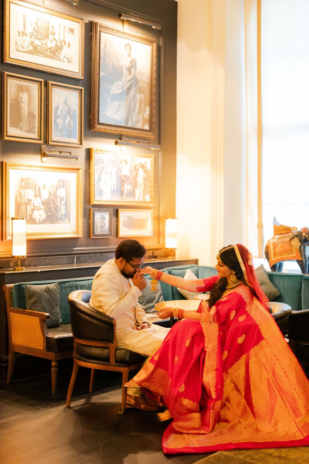 Indian couple in traditional wedding attire sitting together in an elegant room with vintage framed photos on the wall.