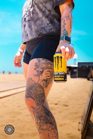 Tattooed woman holding a Monster Rehab energy drink can on a sunny beach boardwalk.