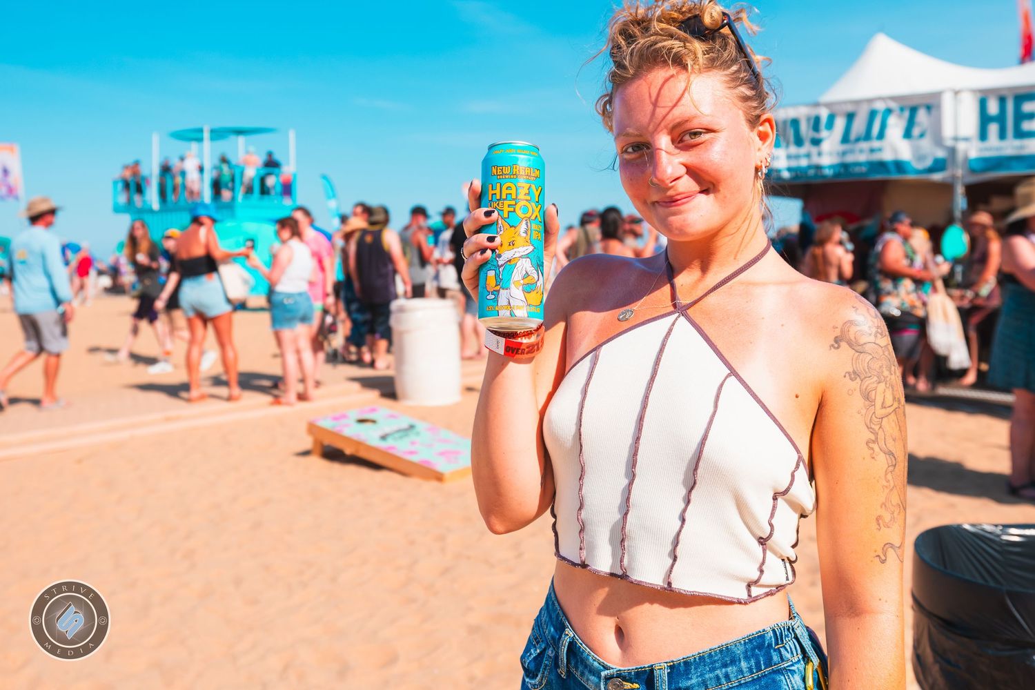Man in tropical shirt raising two beer cans at sunny Virginia beach music festival.