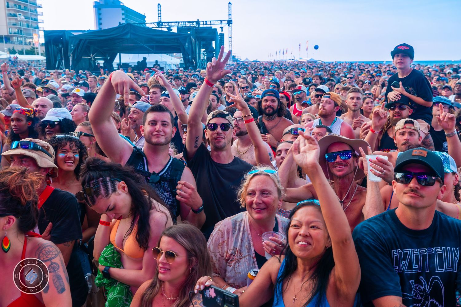 Large crowd of enthusiastic fans at outdoor music festival enjoying live performance on sunny day.