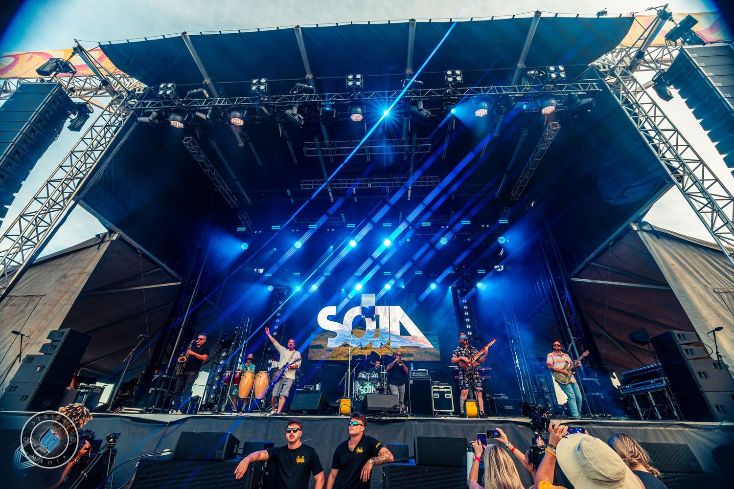 SOJA band performing on large outdoor festival stage with blue lighting and tall speaker towers.