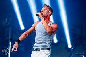 Muscular male vocalist in gray tank top singing passionately on stage with blue concert lighting.