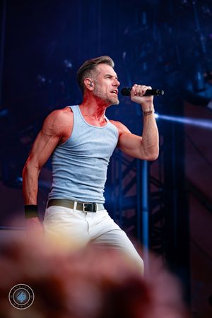 Athletic male singer in gray tank top and white pants performing energetically at outdoor concert.