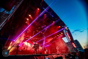 Outdoor concert stage with vibrant purple laser lights and red fog during evening performance.