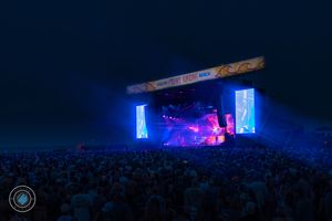 Massive outdoor music festival crowd watching illuminated stage with blue lighting in evening sky.