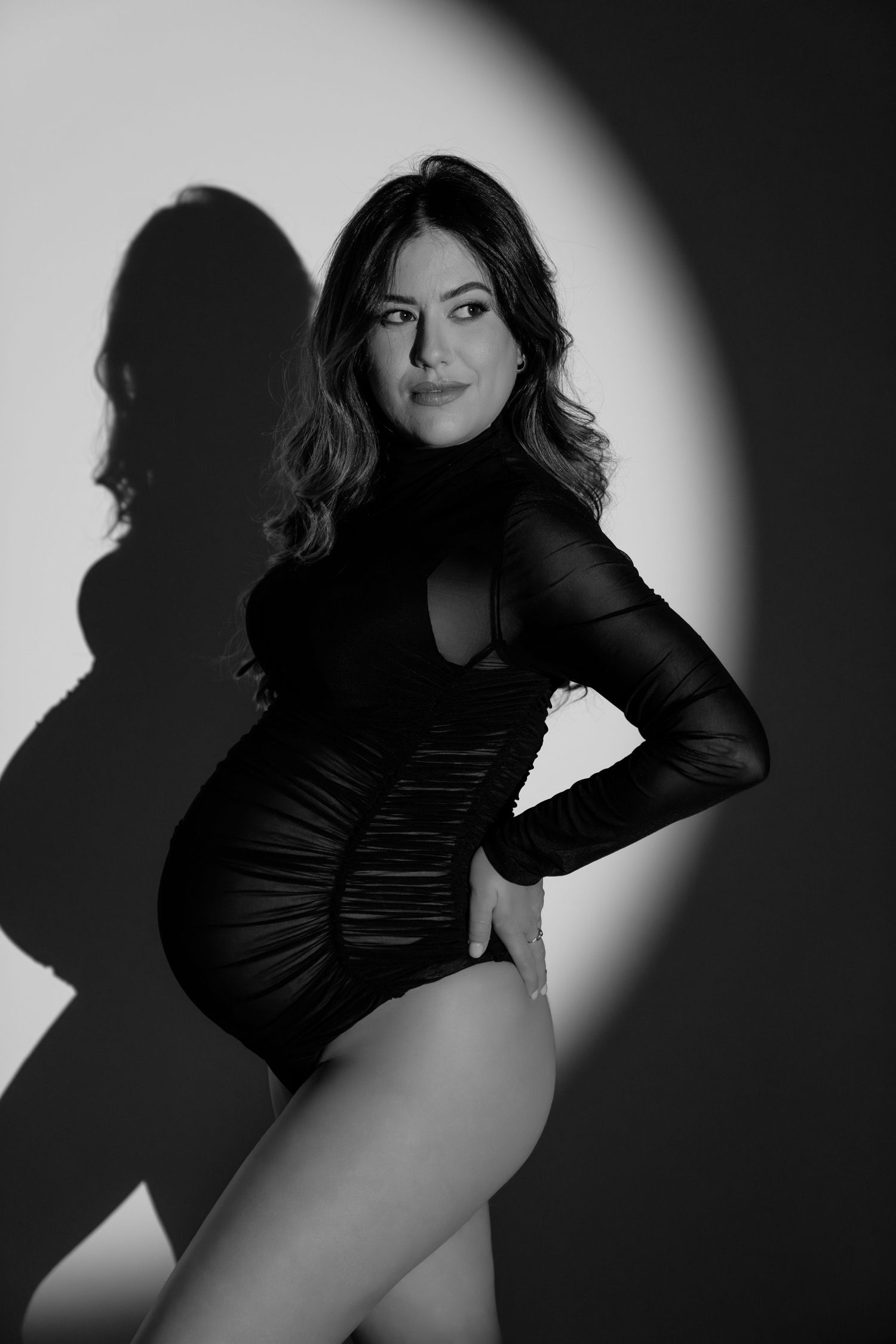 Black and white maternity photo of a pregnant woman in a black bodysuit posing gracefully in a spotlight.