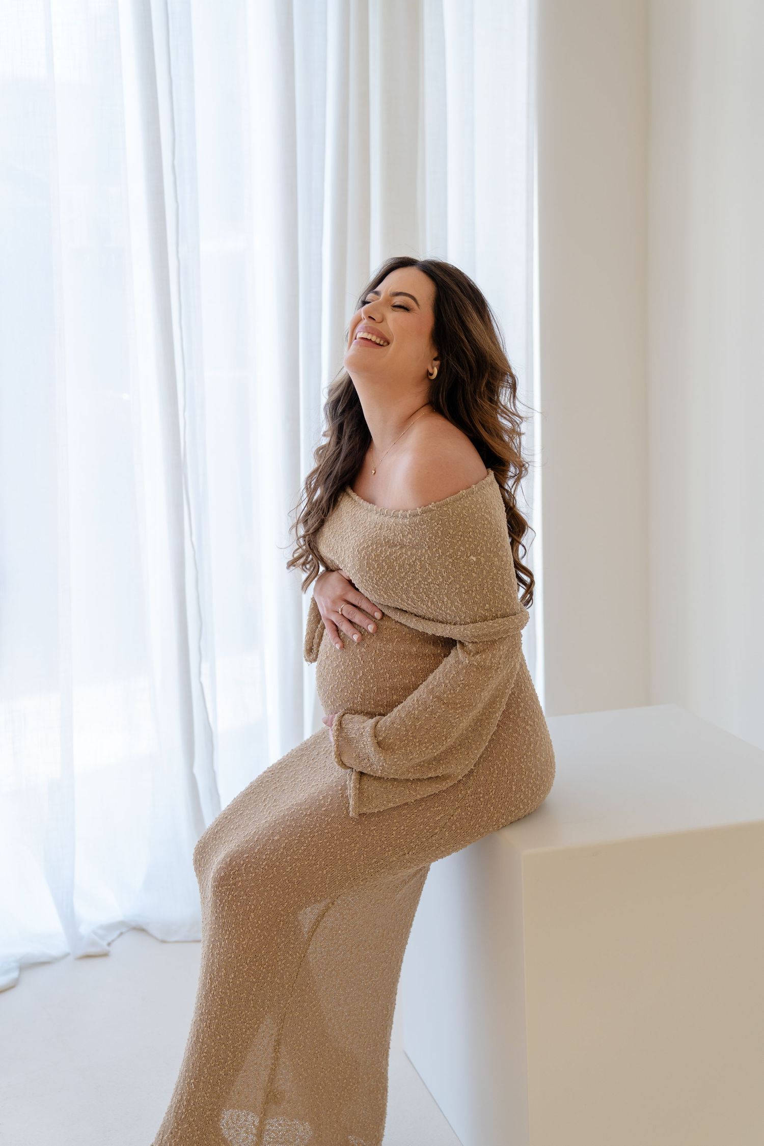 Pregnant woman in beige knit off-shoulder gown sitting by window, smiling joyfully while cradling her baby bump.