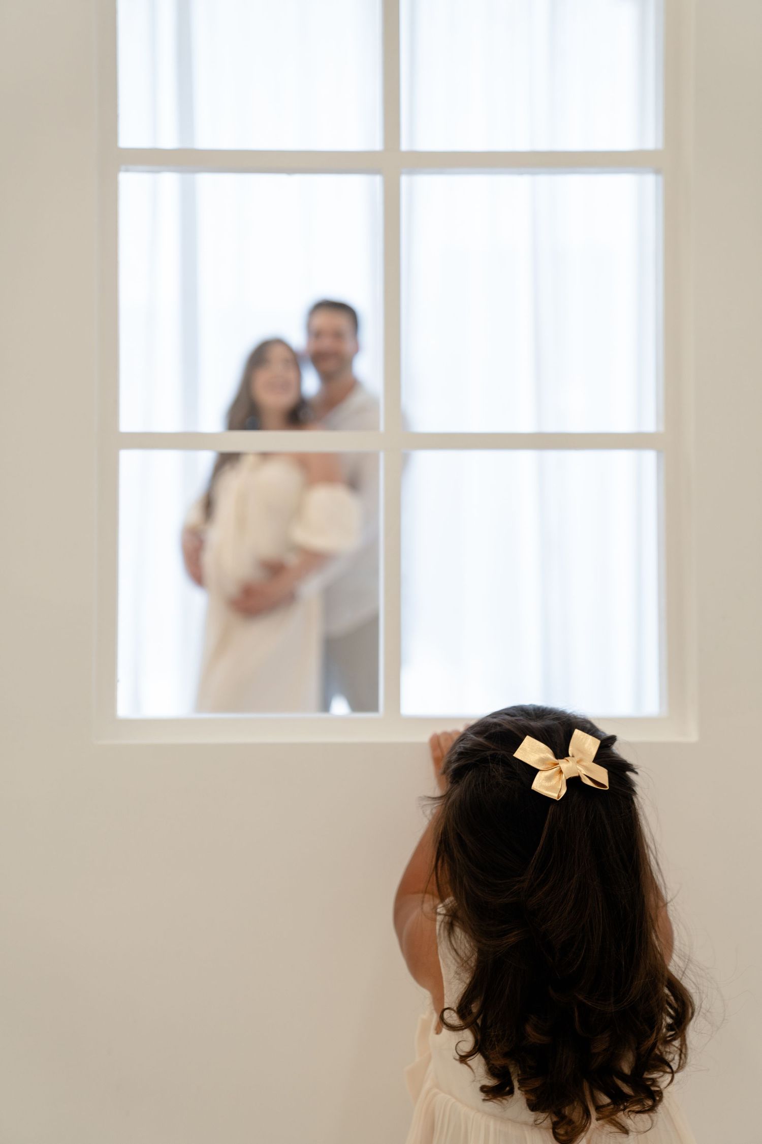A young girl with a bow in her hair looks through a window at a couple embracing, holding a newborn baby.