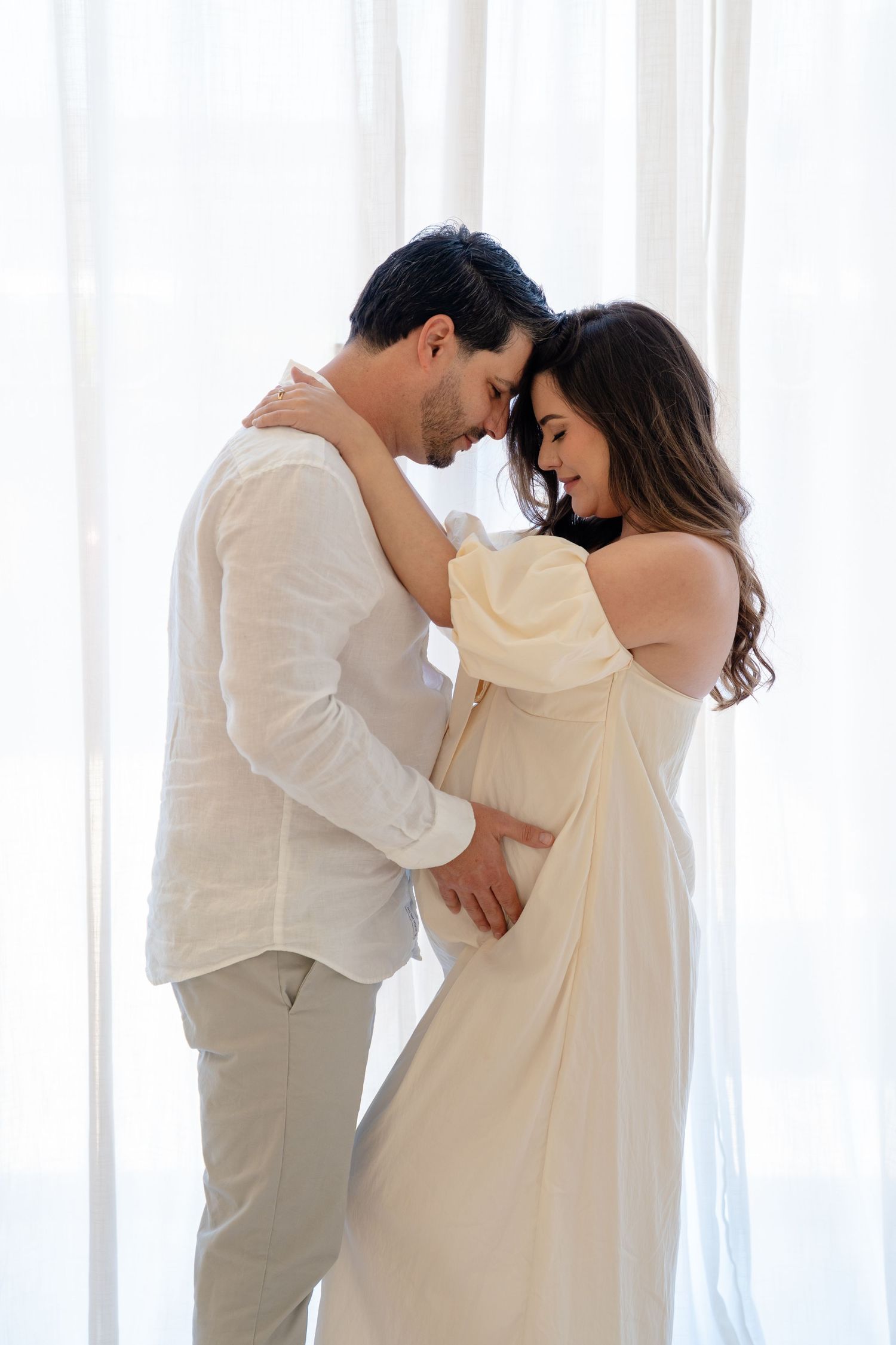 A couple in white clothing shares an intimate moment, with the woman in a flowing dress resting her forehead against her partner.