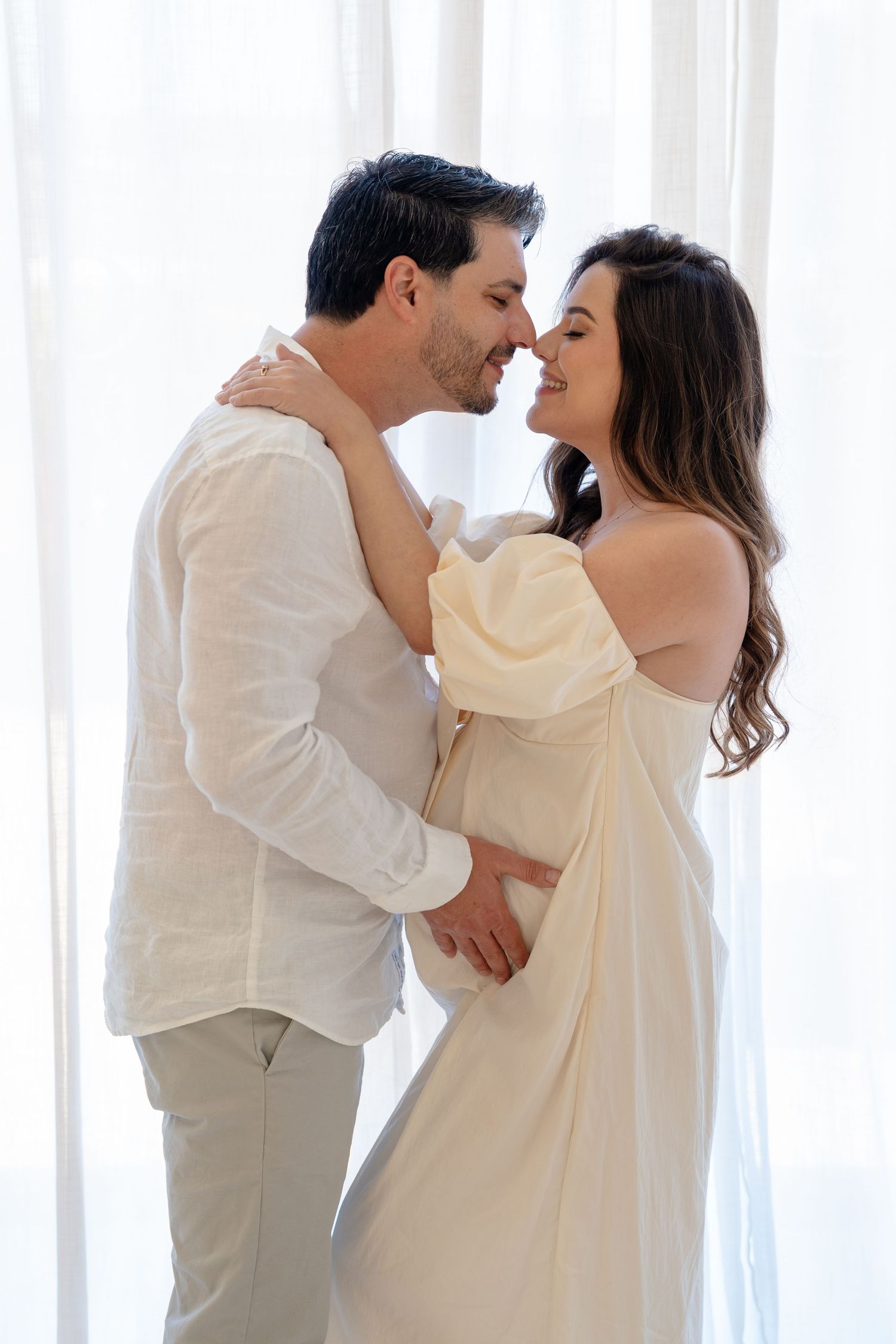 A couple in white outfits share a romantic moment, with the woman in an off-shoulder dress and the man smiling tenderly.