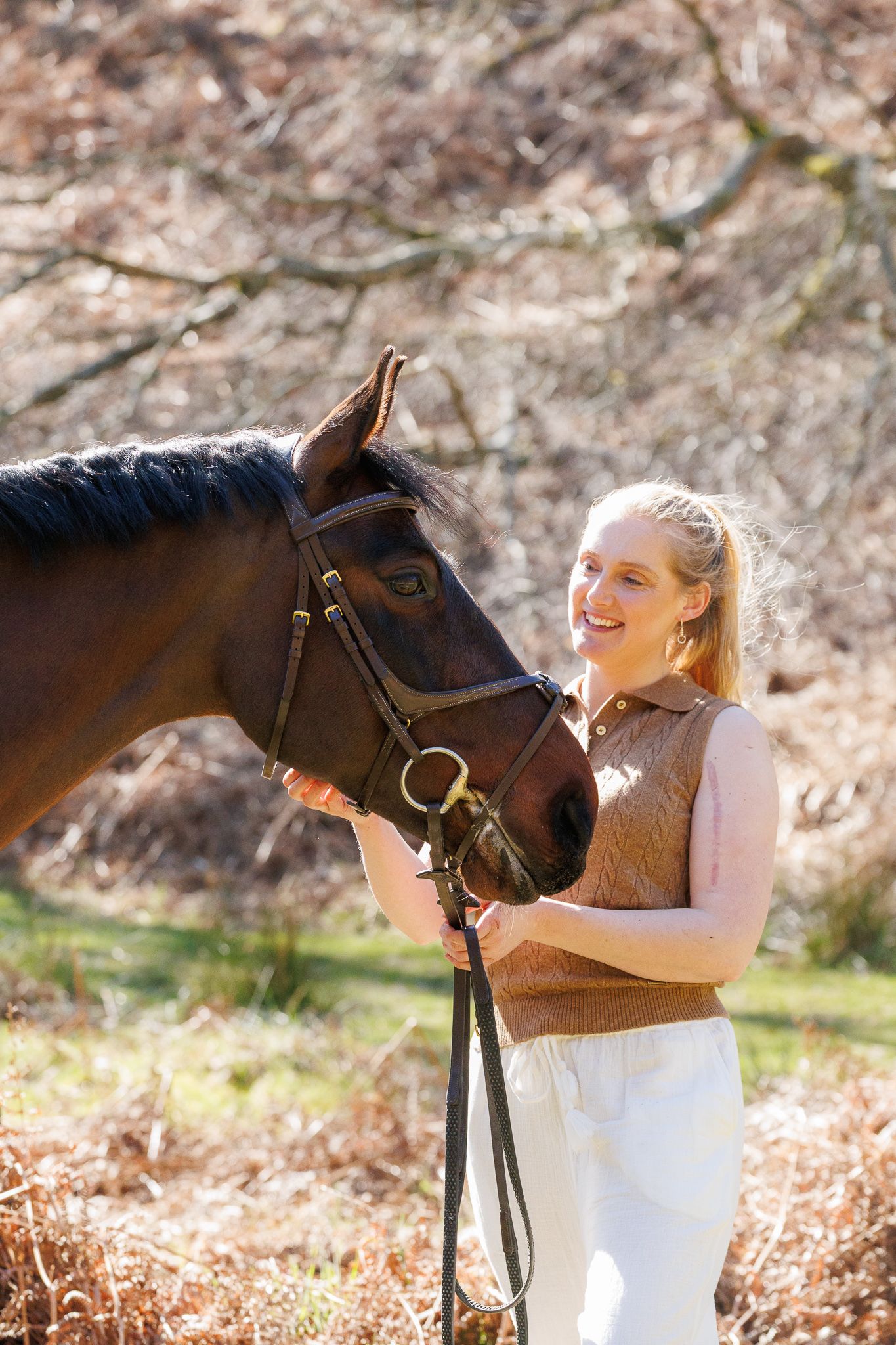 Smiling blonde woman in brown vest holding reins of a bay horse outdoors in a sunny woodland setting.