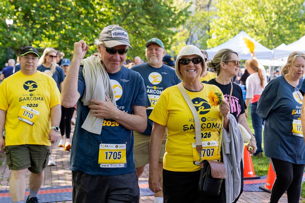 Participants wearing yellow Sarcoma race bibs celebrate at an outdoor charity fun run event on a sunny day.