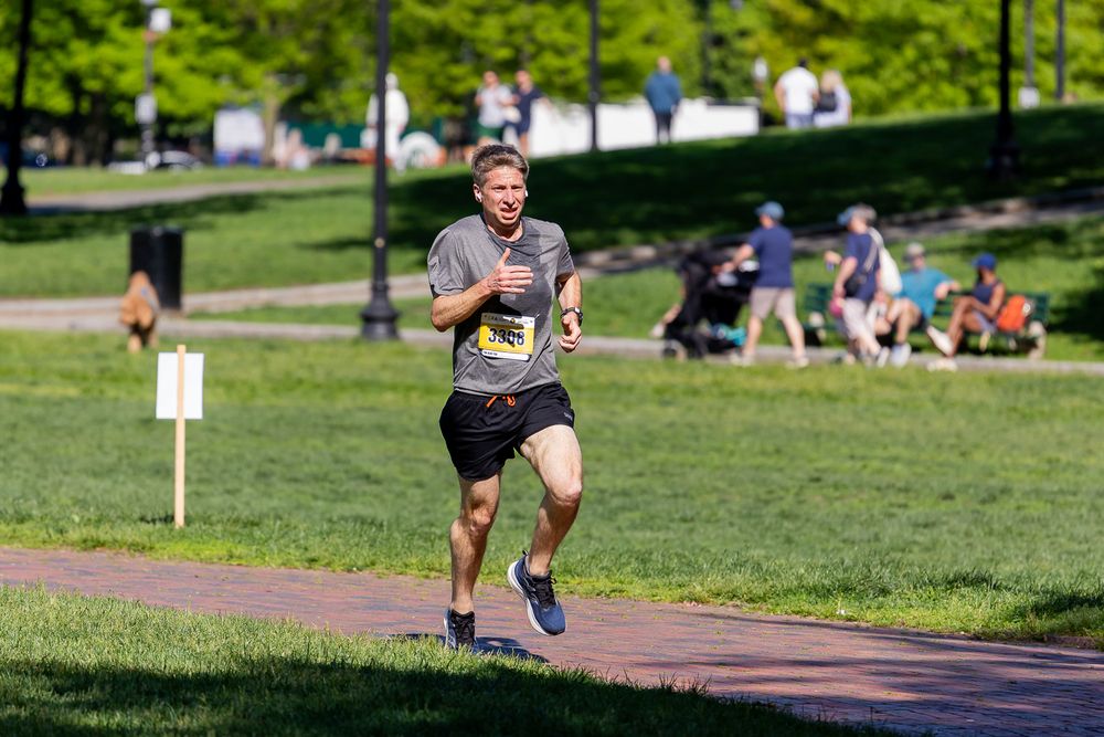 Man running on a park path during a race on a sunny day with green grass and trees in the background.