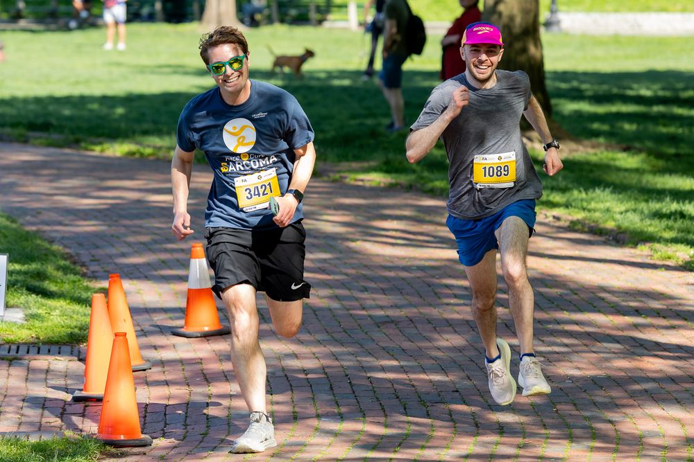 Two runners competing in the Sarcoma Foundation race on a brick path through a sunny green park.